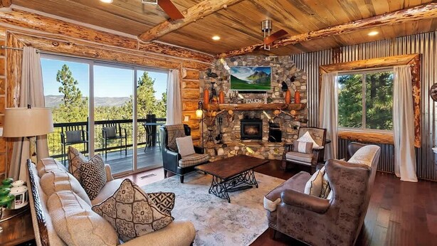 A log cabin interior with a stone fireplace, wooden ceiling, and large windows offering a mountain view. - Designer Chalet | Sauna · Fireplace · Mountain Views (Big Bear Lake)