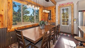 A wooden dining room with a large table and chairs, a chandelier, and a view of trees through large windows. - Designer Chalet | Sauna · Fireplace · Mountain Views (Big Bear Lake)