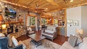 A cozy living room with a stone fireplace, wooden ceiling beams, and a large window. - Designer Chalet | Sauna · Fireplace · Mountain Views (Big Bear Lake)