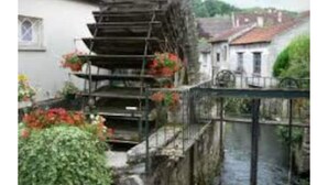 Un moulin à eau traditionnel, doté d’un escalier menant à une plateforme, entouré de verdure et d’un petit bâtiment.