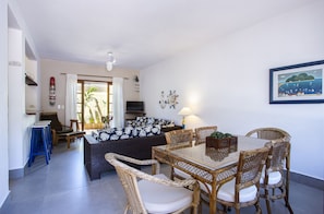 A dining area with a glass-top table and wicker chairs, a sofa with patterned cushions, and a television in the background. - Residencial Santa Lucia (São Sebastião)