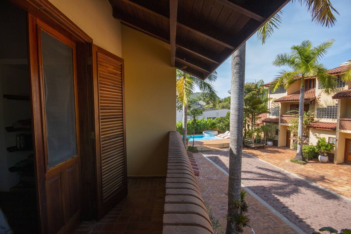 A covered balcony with a bench, a pool, and a view of a residential area with palm trees and houses.