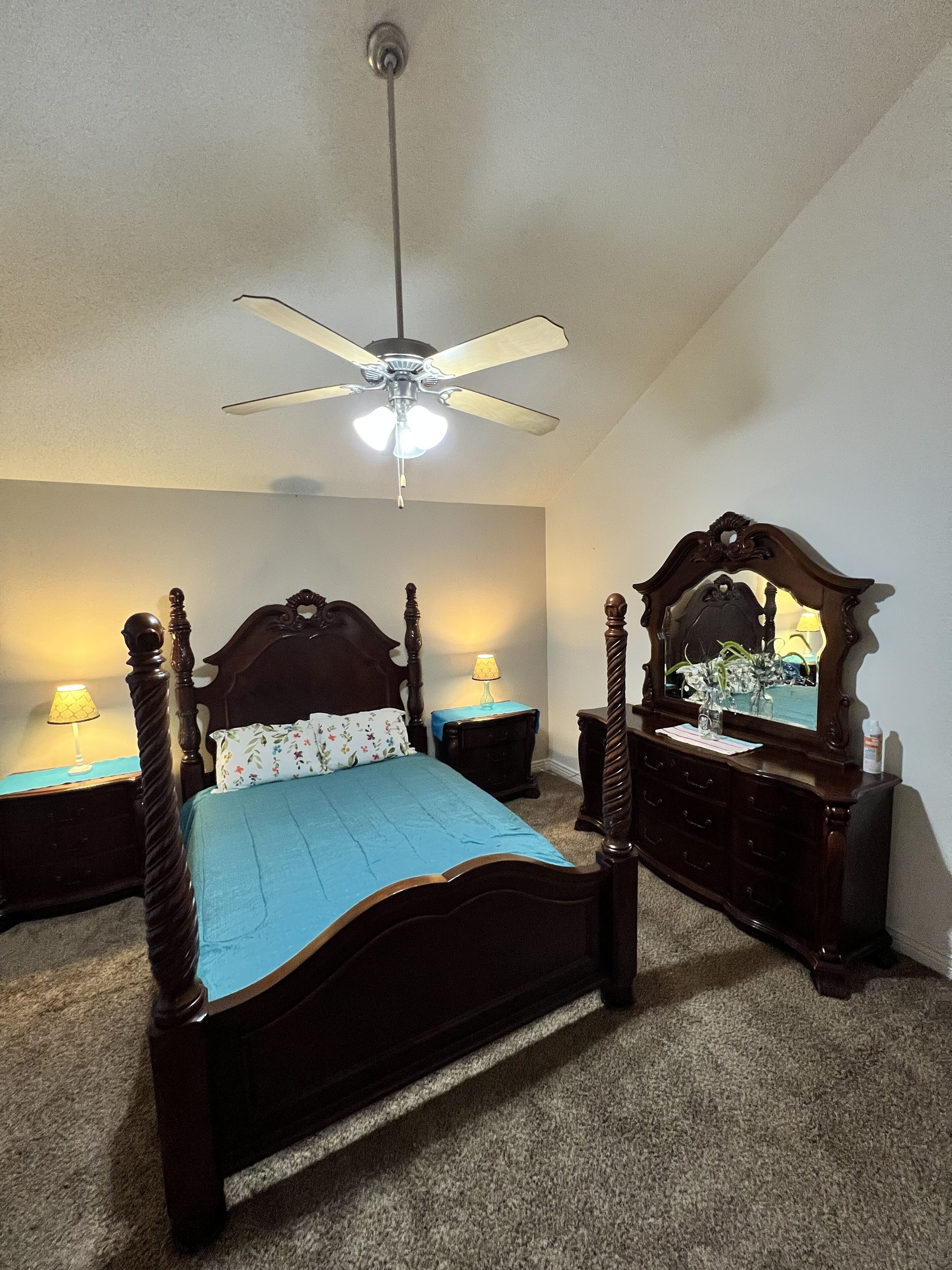 Une chambre à coucher avec un lit à baldaquin, un ventilateur de plafond, une table de chevet avec une lampe, une commode avec un miroir et un sol recouvert d’une moquette.