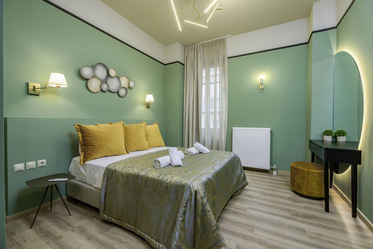 A neatly arranged bedroom with a bed, bedside lamps, a wall art piece, a window with curtains, and a small table with a lamp.