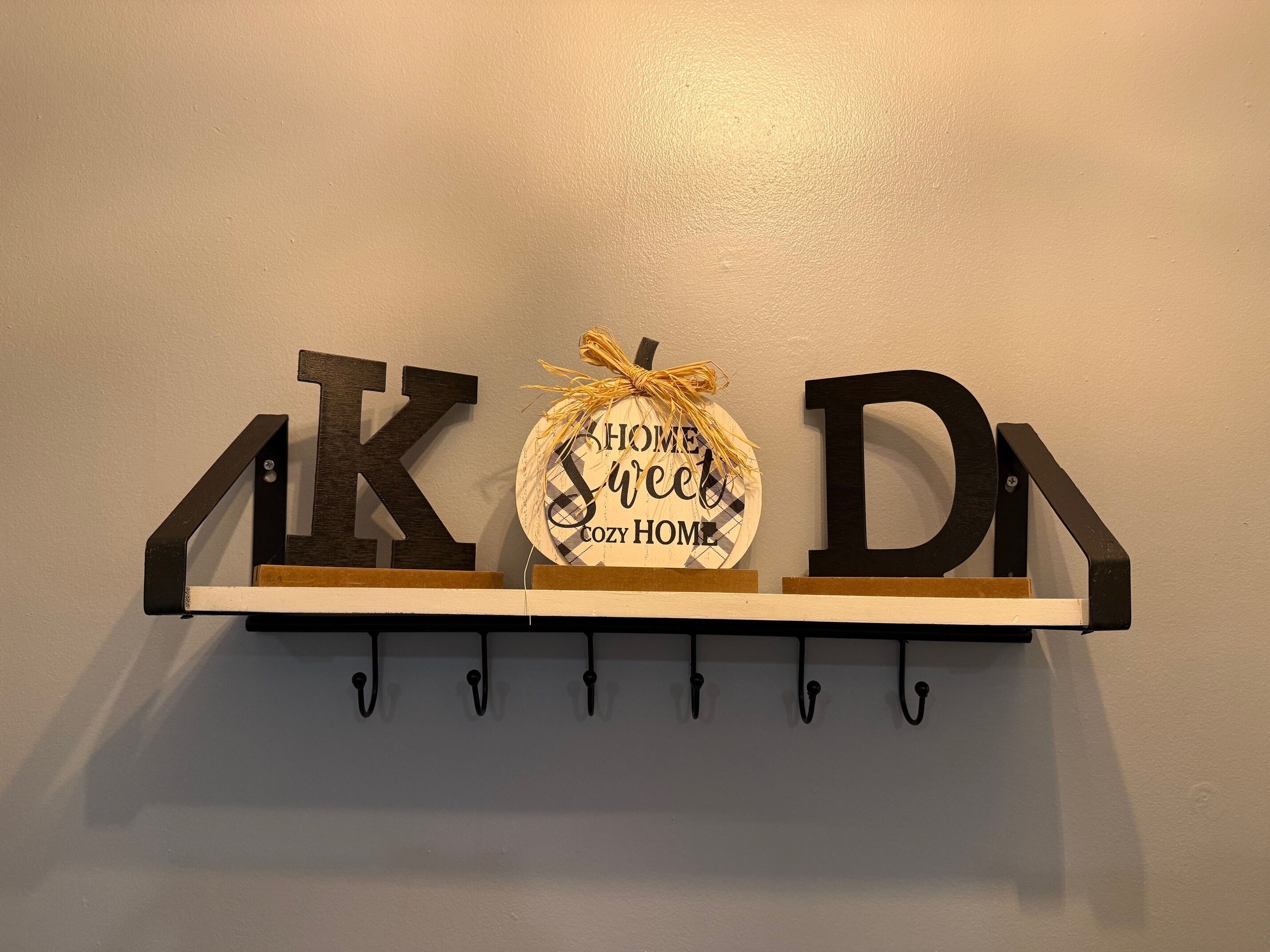 Uma prateleira na parede com três letras de madeira formando a palavra "KOD" e um sinal circular com a frase "CASA ACONCHEGANTE". 