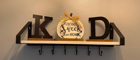 Uma prateleira na parede com três letras de madeira formando a palavra "KOD" e um sinal circular com a frase "CASA ACONCHEGANTE".