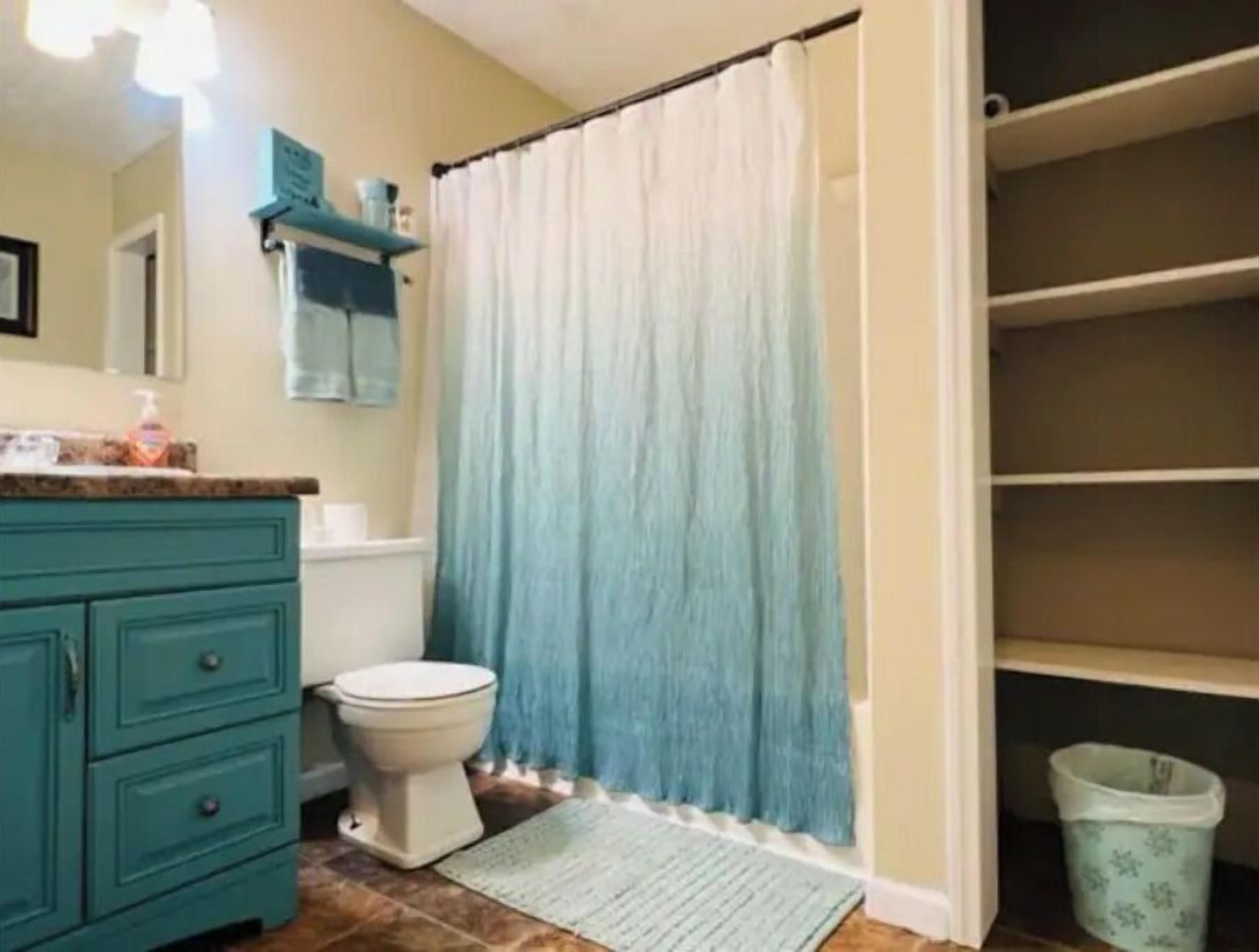 Un baño con tocador color turquesa, inodoro blanco, cortina de ducha azul y un cubo de basura blanco.