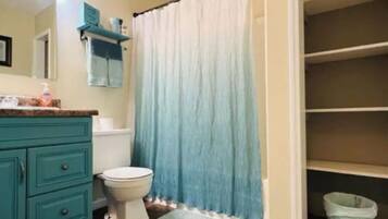 Un baño con tocador color turquesa, inodoro blanco, cortina de ducha azul y un cubo de basura blanco.