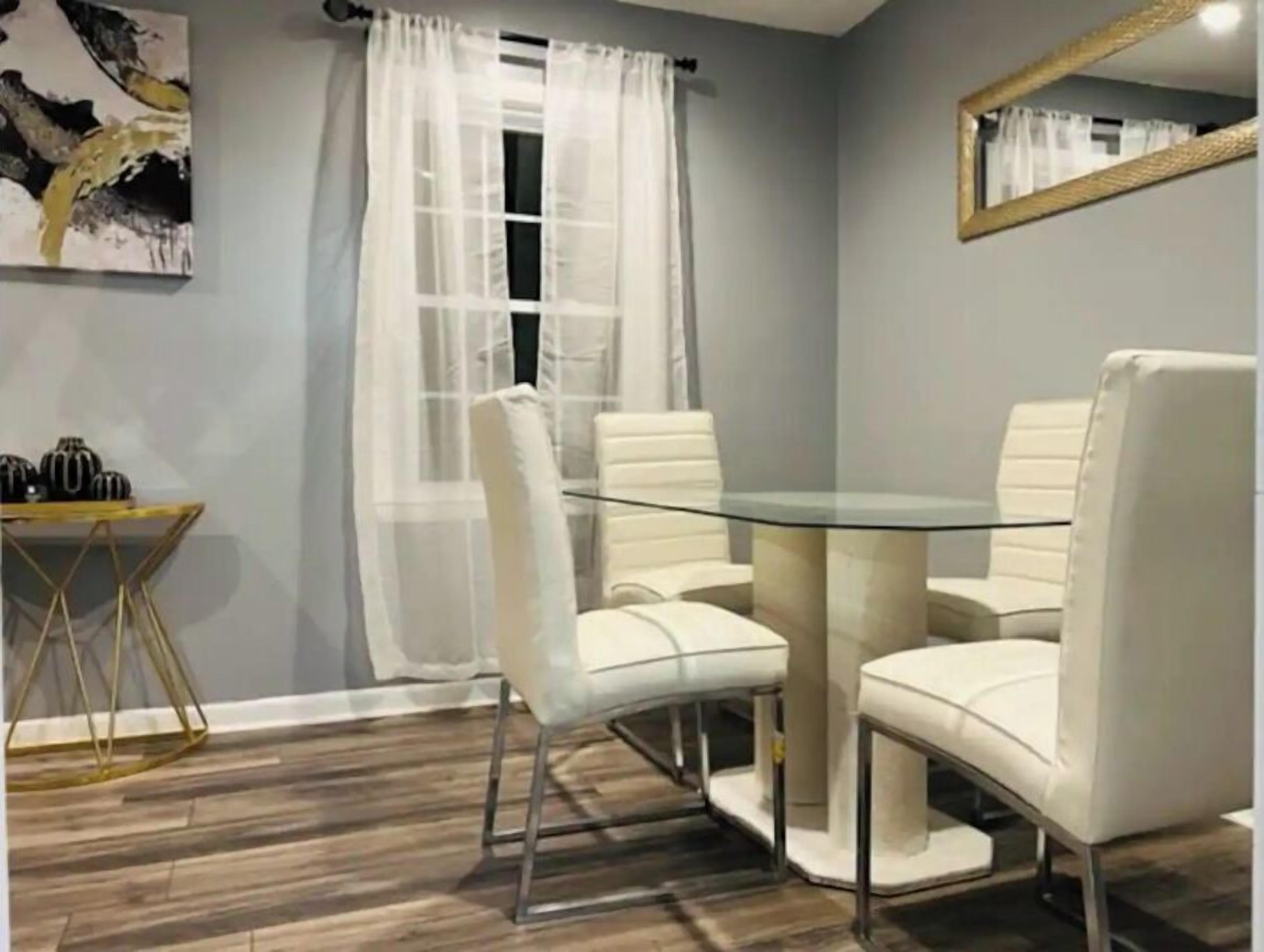 Un coin repas avec une table en verre et des chaises blanches, un plancher en bois, un tableau au mur et une fenêtre ornée de rideaux légers.