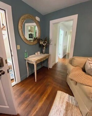 Une pièce avec un plancher en bois, un canapé beige, une console blanche avec un miroir et une porte donnant sur une autre pièce.