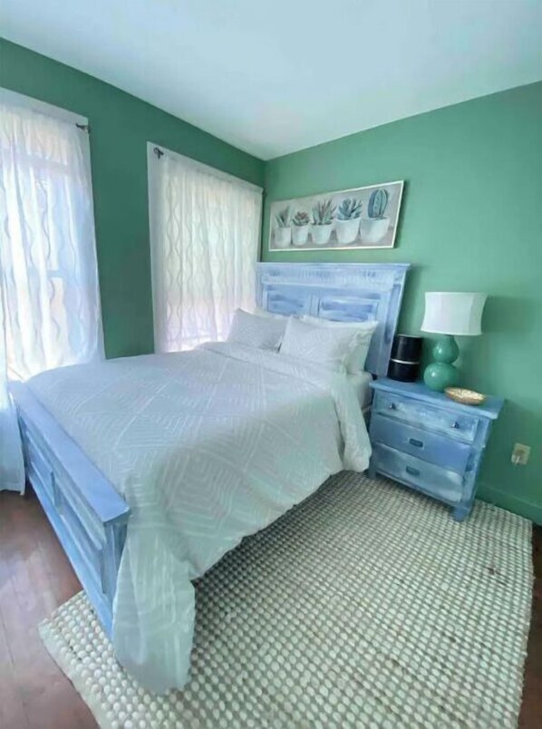 Une chambre à coucher avec un lit, une table de chevet, une lampe et un mur orné d’un tableau représentant une plante.