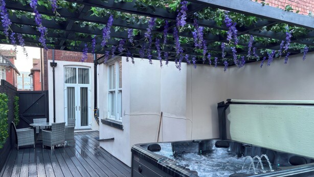 A patio with a hot tub, a pergola with hanging purple flowers, and a seating area with chairs and a table. - Orchard Lodge (Lytham St. Anne's)