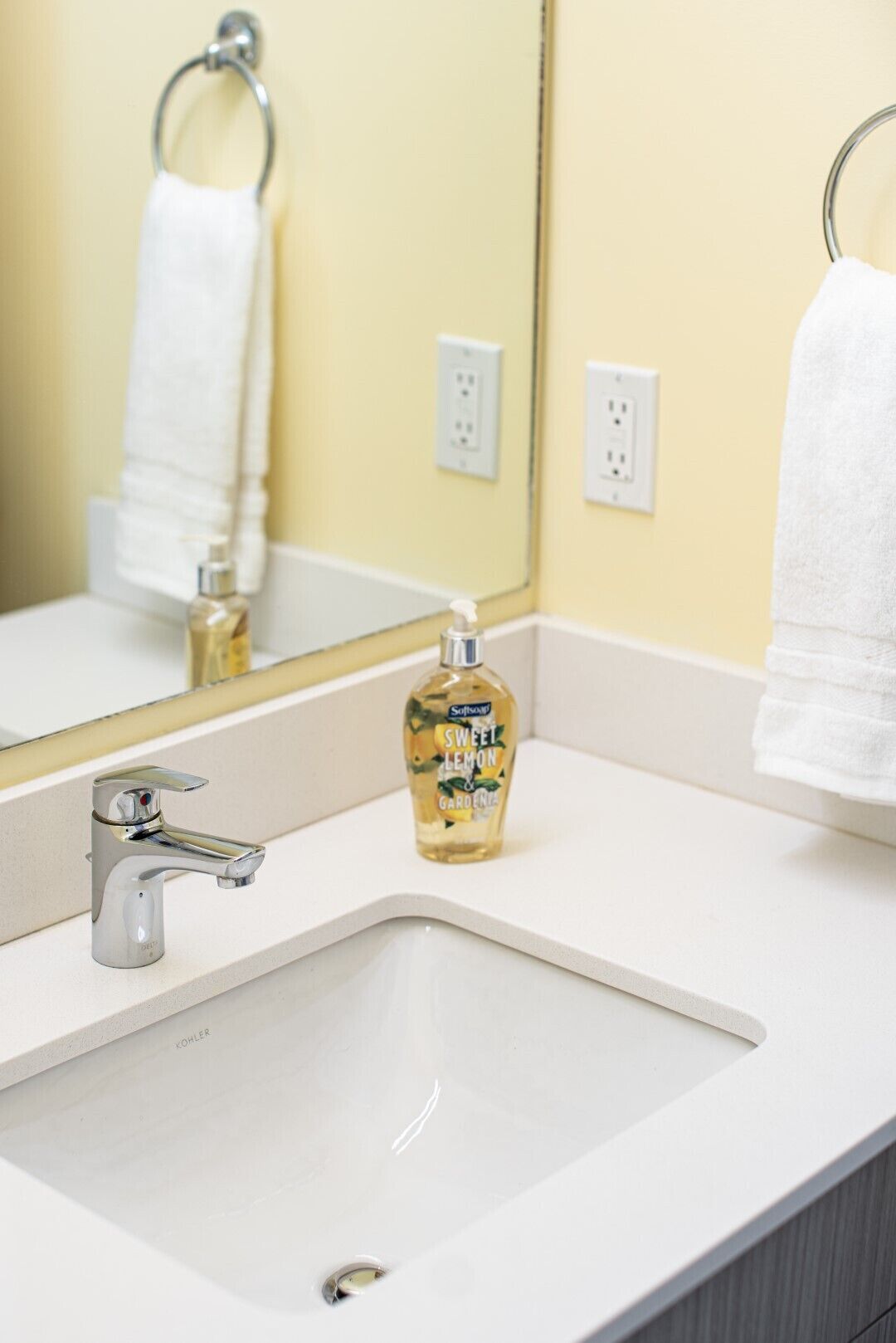 A bathroom sink with a chrome faucet, a bottle of Softsoap Sweet Lemon Gardenia hand soap, and a white towel hanging on a metal towel ring.