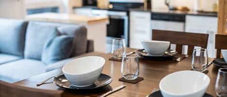 A dining table set with white bowls, clear glasses, and cutlery, with a modern kitchen in the background.