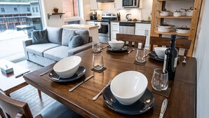 A dining area with a wooden table set for four, including bowls, plates, and cutlery. A wine bottle and glasses are also visible. The room features a grey sofa, a kitchen in the background, and a view of buildings outside.