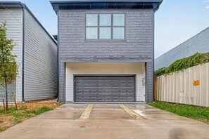 Une maison à deux étages avec un portail de garage, une façade en pierre et un petit jardin devant, agrémenté d’un arbre et d’une clôture.