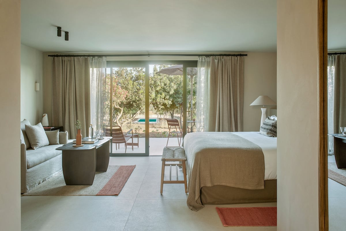 A hotel room with a bed, a sofa, a coffee table, and a dining table. There is a large window with curtains, and a view of the outdoors.