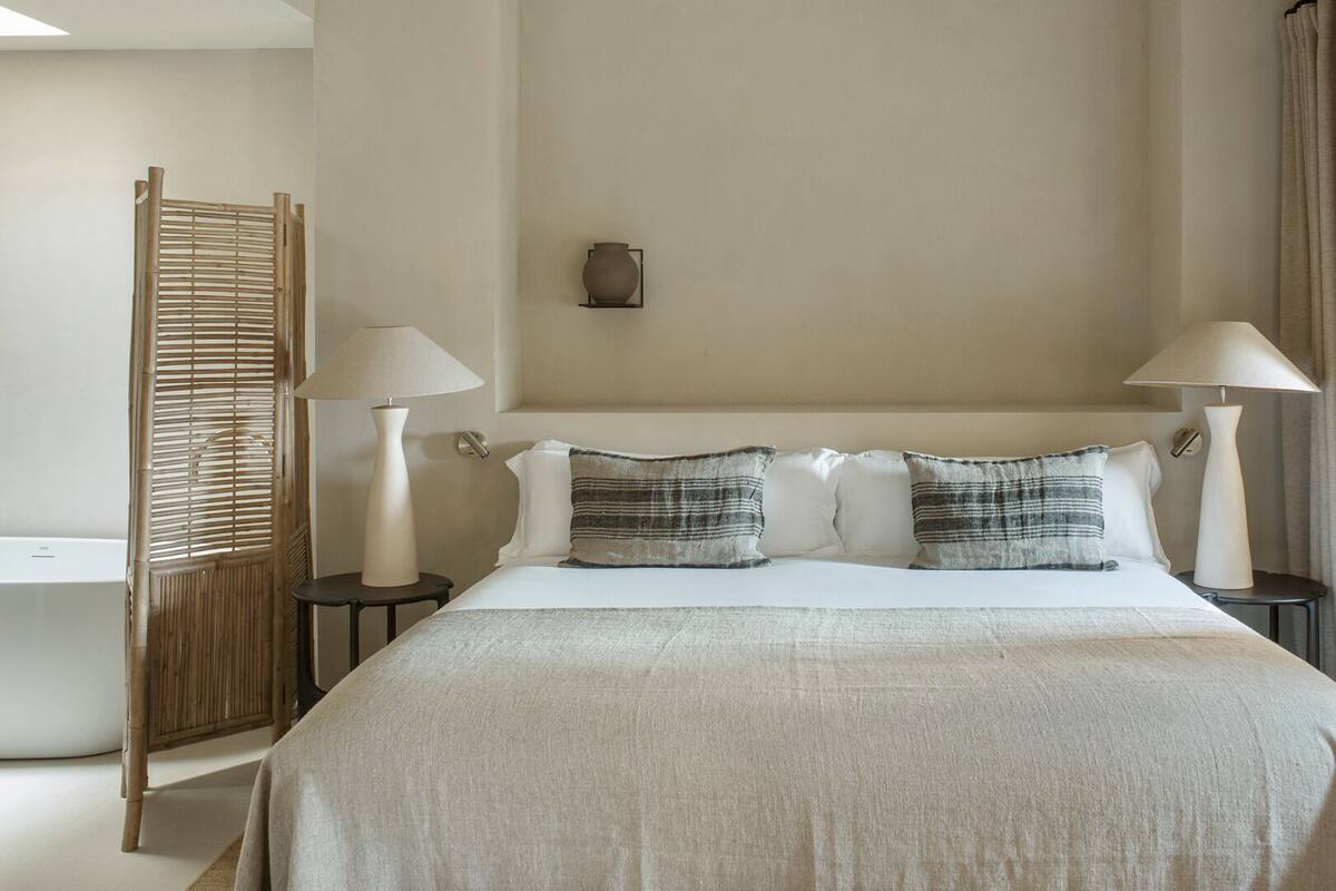 A hotel room with a bed, two bedside tables, two lamps, a wall-mounted light fixture, and a large window with curtains.