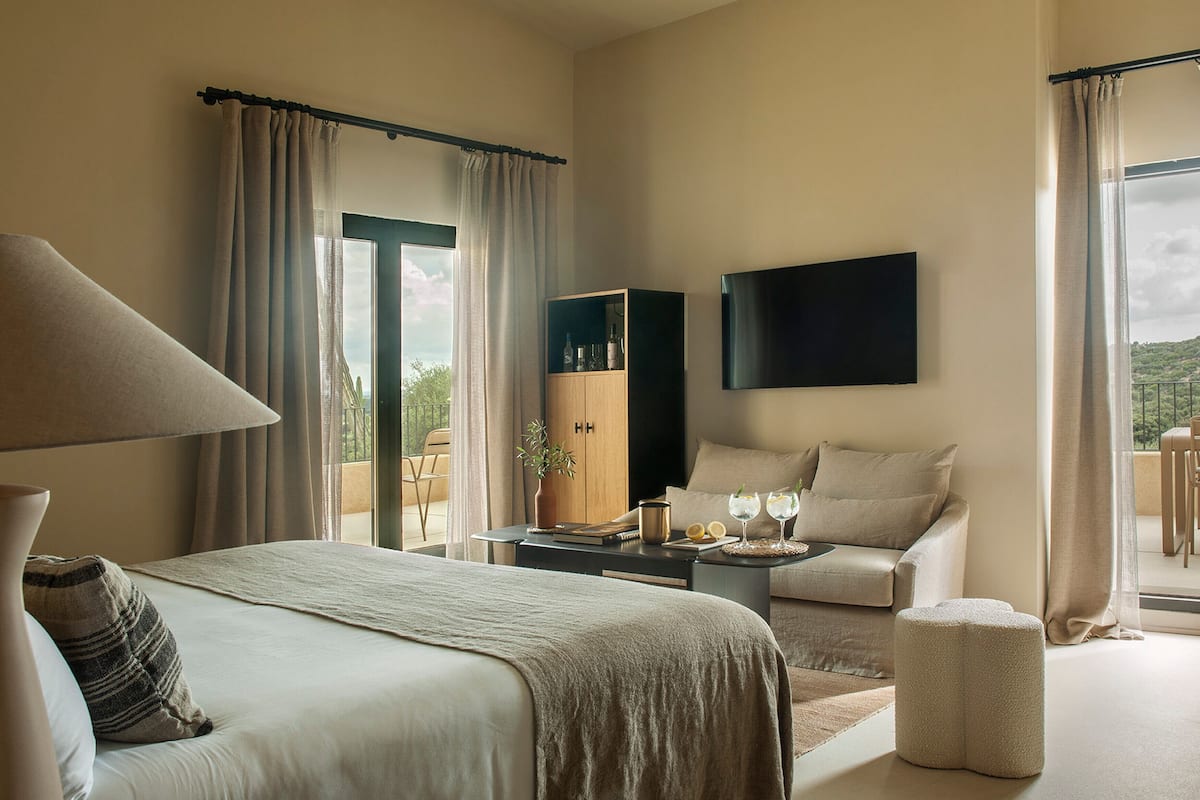 A modern hotel room with a bed, a desk, a chair, a TV, and a balcony with curtains.