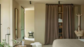 Habitació d'hotel amb un llit, una taula de nit amb una copa i ampolla de vi, un mirall, una estantària amb tovalloles i una cistella penjant.