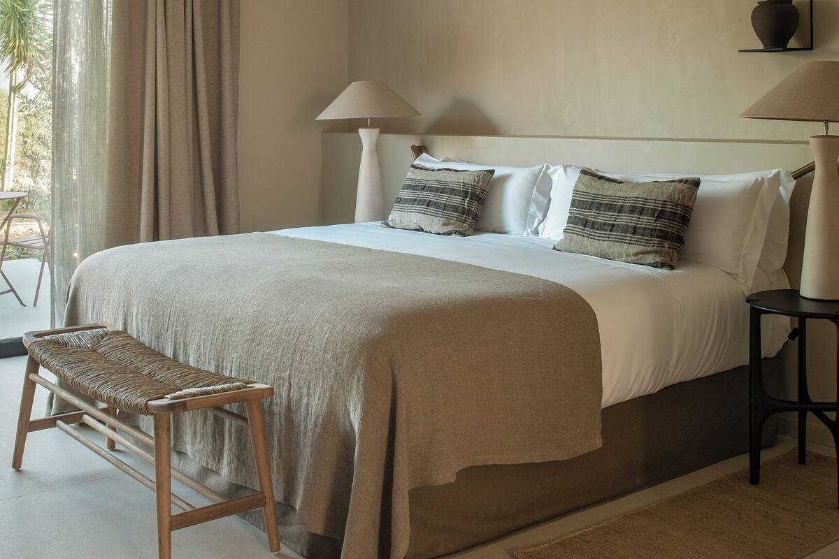 A bedroom with a bed, two bedside lamps, a bench, and a view of the outdoors.