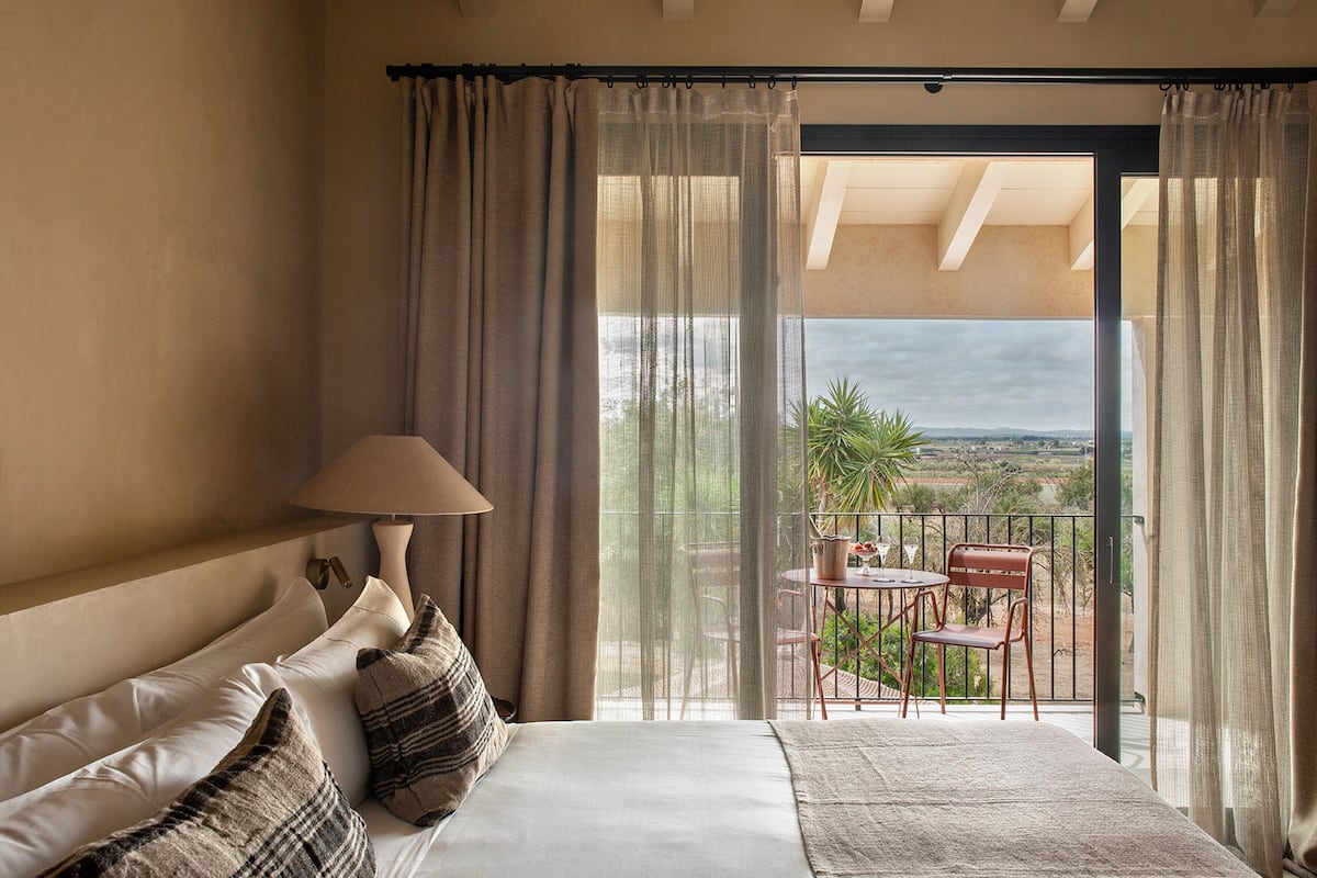 A bedroom with a bed, a nightstand, a lamp, and a view of an outdoor balcony with a table and chairs.