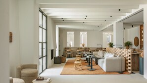 Un amplio salón con un sofá grande, una mesa de centro y una mesa de comedor. Hay sillas de mimbre y una pequeña mesa redonda. La habitación tiene techos altos con vigas a la vista y ventanas grandes.