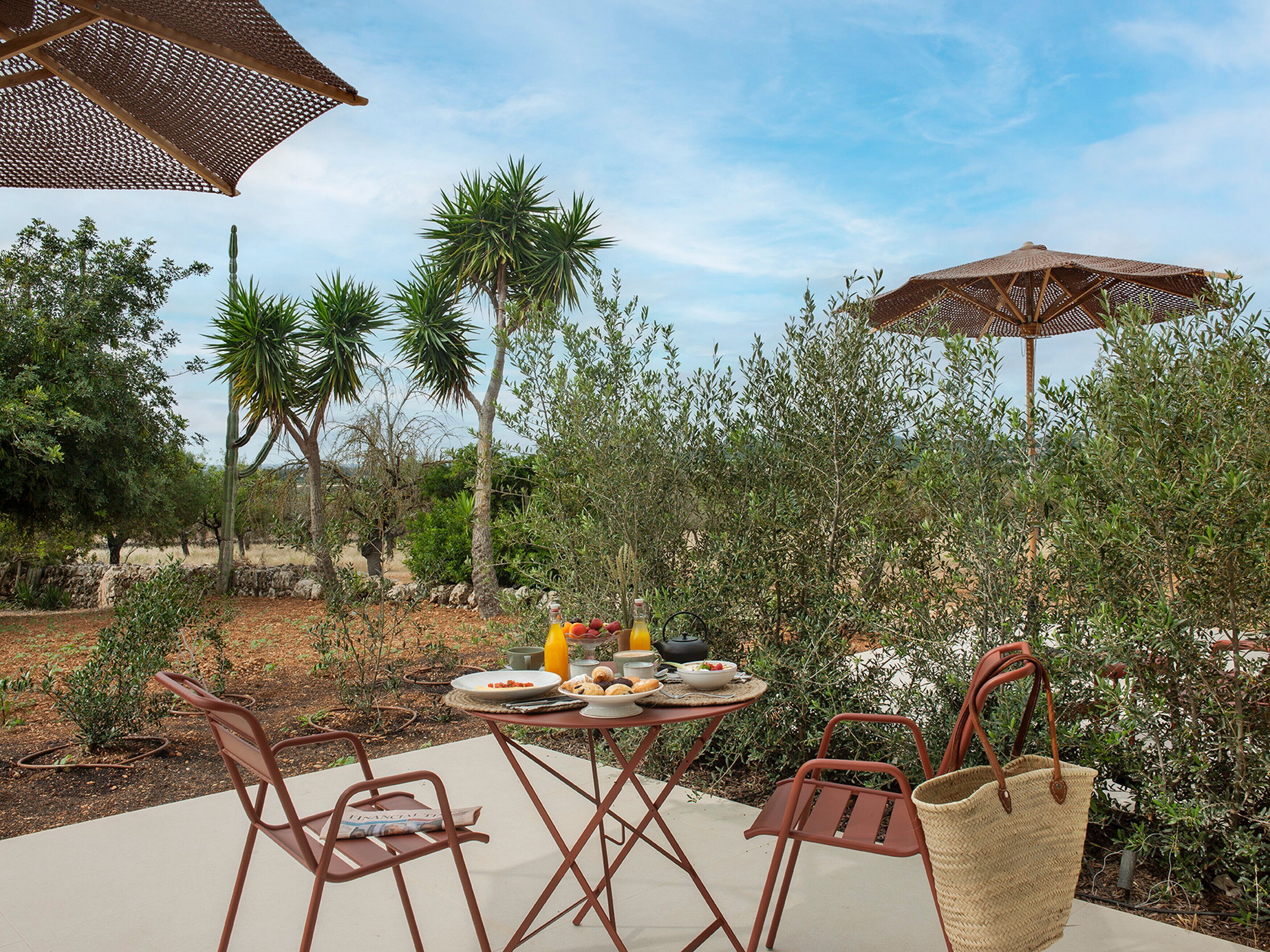 Un área de comedor al aire libre con una mesa para dos, rodeada de sillas y sombrillas, en un jardín con diversas plantas y árboles.