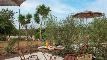 Un área de comedor al aire libre con una mesa para dos, rodeada de sillas y sombrillas, en un jardín con diversas plantas y árboles.
