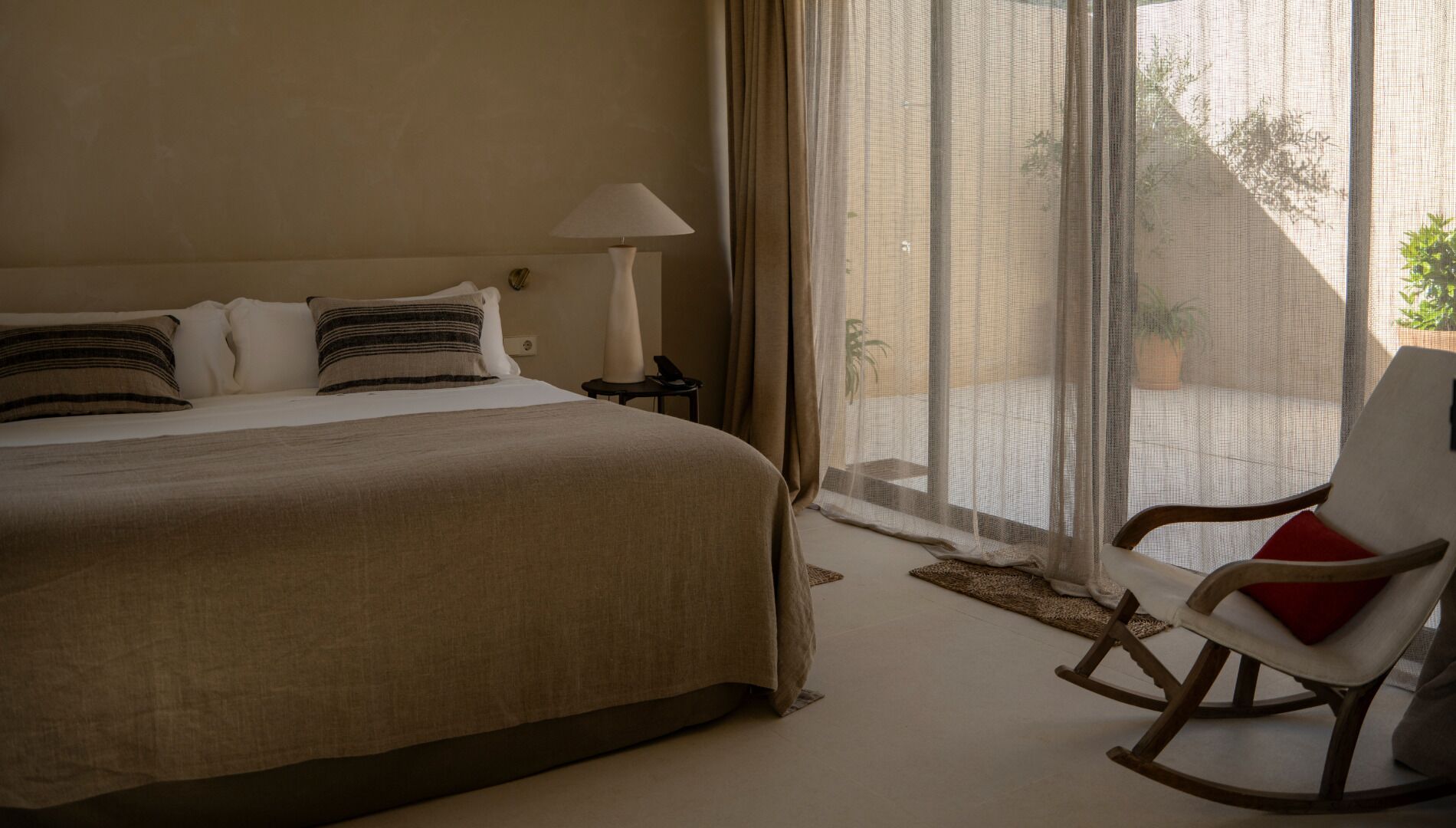 Une chambre avec un grand lit, une chaise, une lampe et une vue sur l’extérieur par une fenêtre.