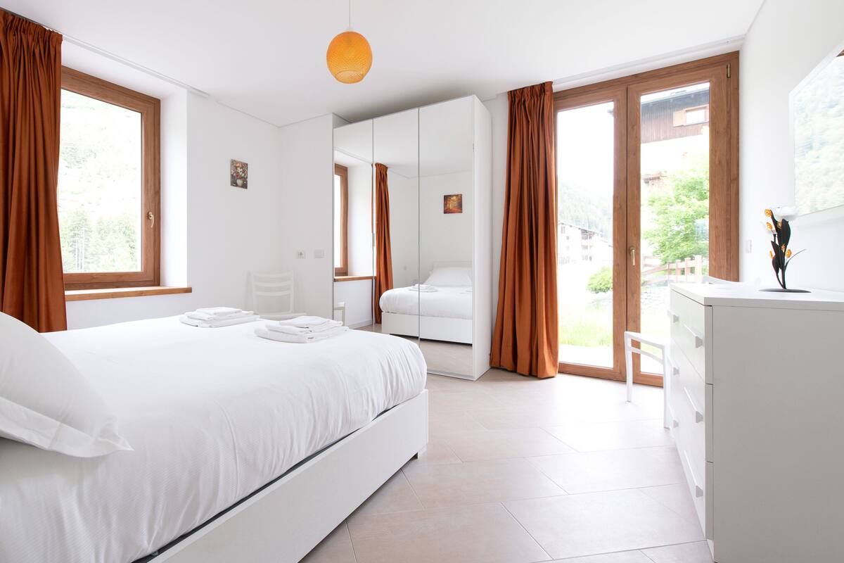 A modern bedroom with a large bed, a wardrobe, a desk, and a view of the outdoors.
