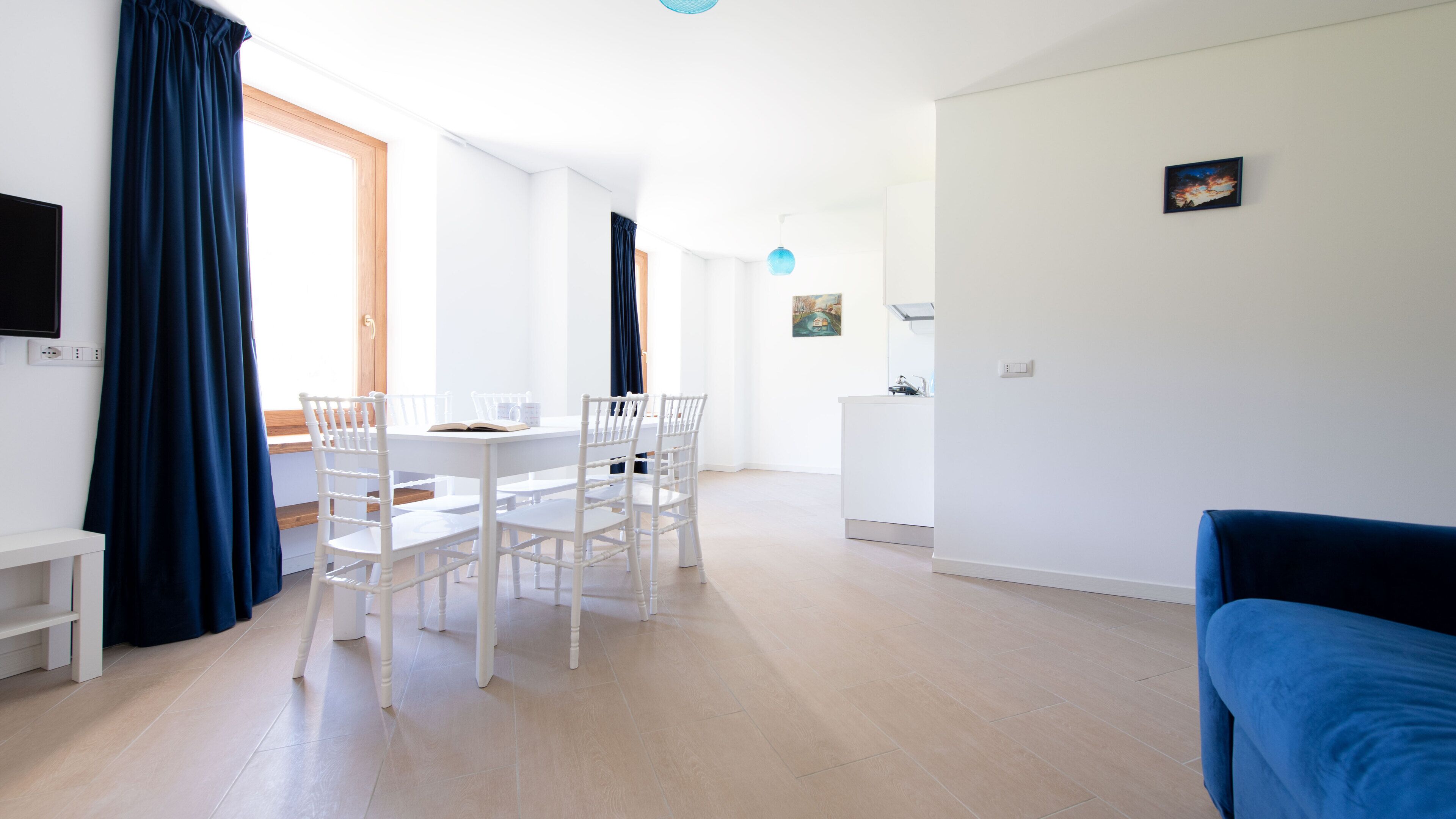 Een moderne woonkamer met een blauwe bank, een eethoek met witte stoelen en een tafel, en een keuken op de achtergrond.