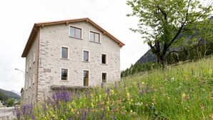 A stone building with multiple windows and a balcony, surrounded by a lush meadow with various wildflowers. - Italianway - Villa Tola (Valdisotto)