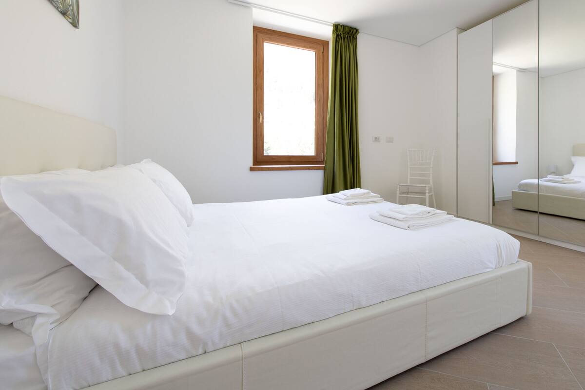 A neatly made bed with white linens and a green curtain, a white chair, and a large mirror reflecting a bed and a window.