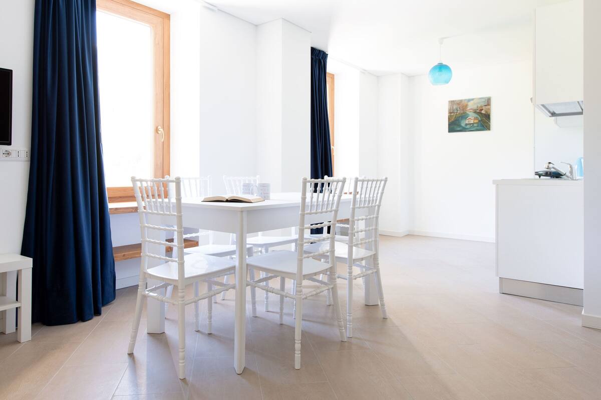 A modern, well-lit room with a dining table, white chairs, a TV, and a kitchen area.