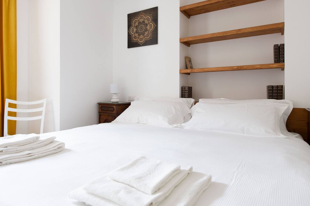 A neatly made bed with white linens, a wooden nightstand with a lamp, and a wall-mounted shelf with books.