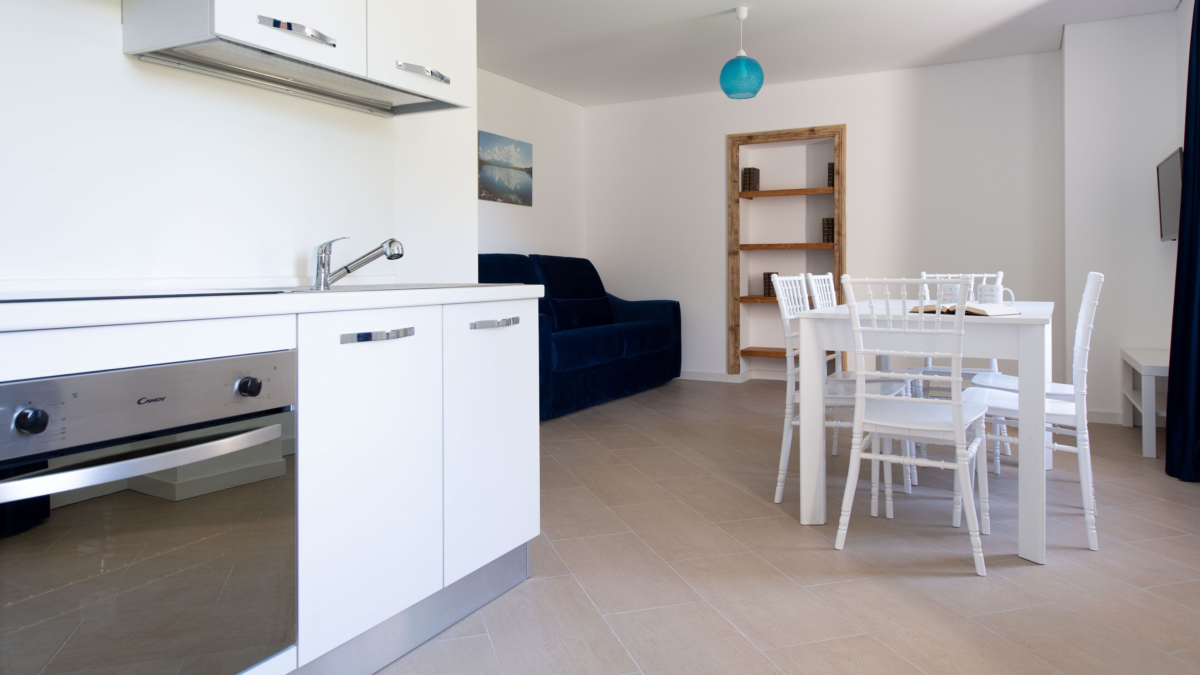A modern kitchen with white cabinetry, a built-in oven, and a sink. There is a dining area with a white table and chairs, and a blue sofa in the background.