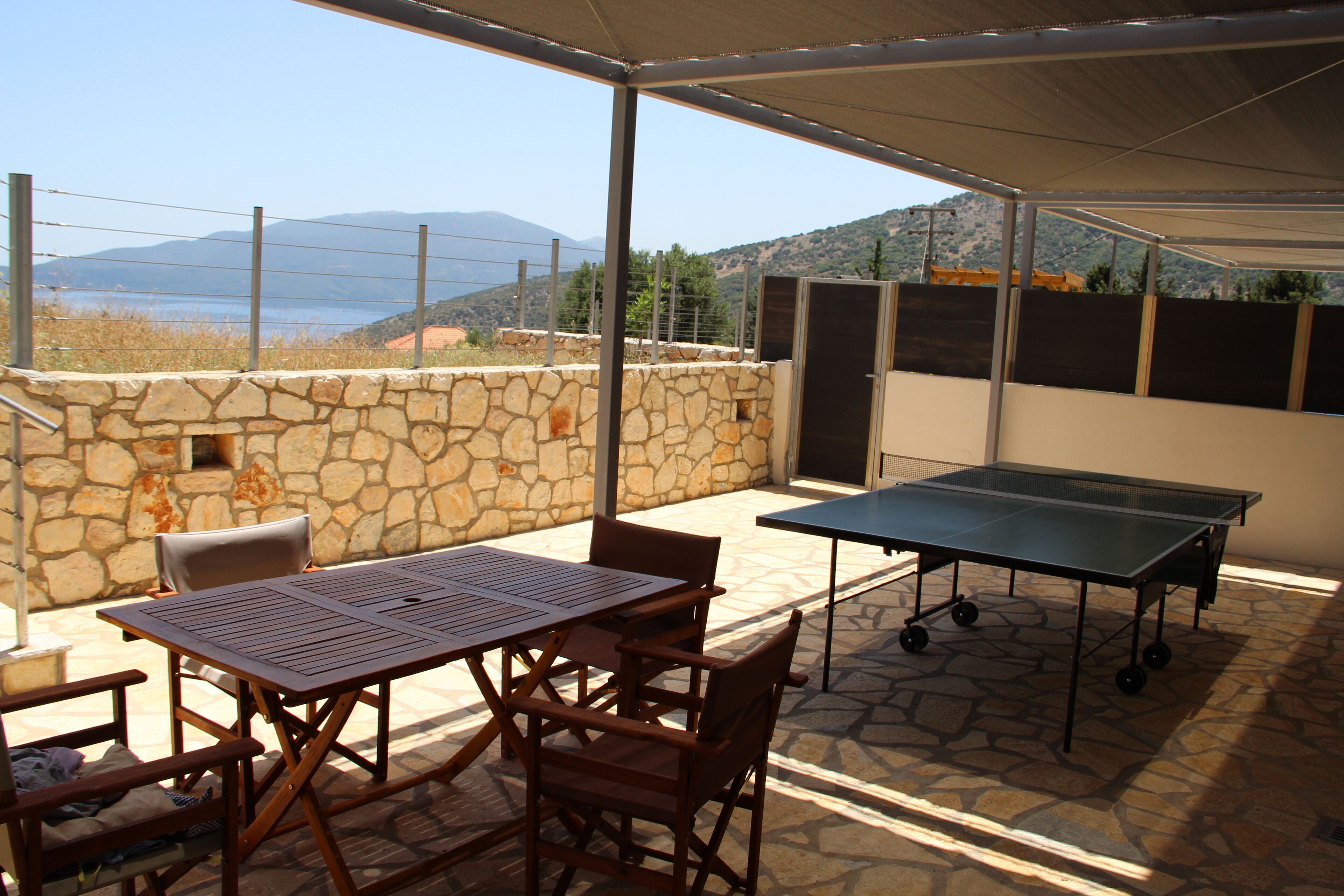 An outdoor patio with a table and chairs, a ping pong table, stone walls, and a view of the sea and mountains.
