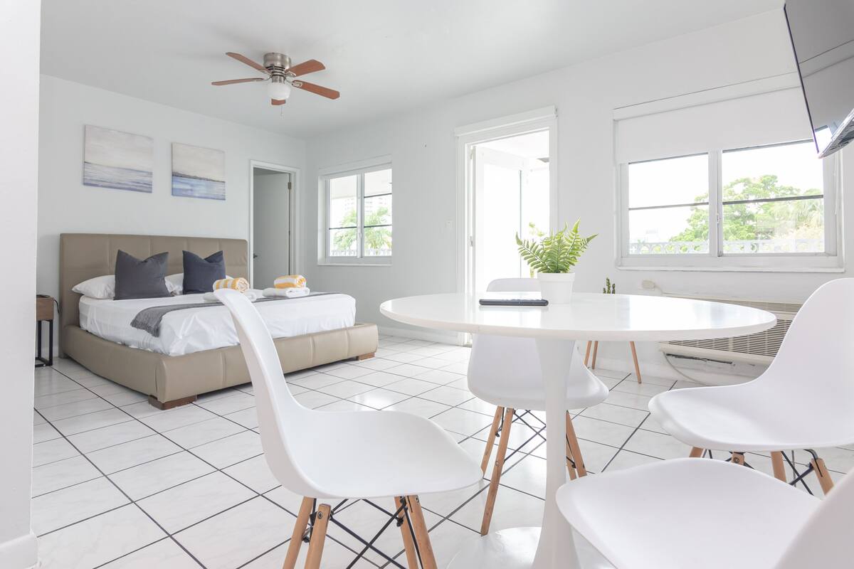 A modern hotel room with a bed, a round dining table, white chairs, a ceiling fan, and large windows.