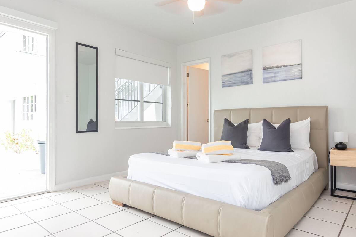 A bedroom with a large bed, a ceiling fan, a window with blinds, and artwork on the wall.