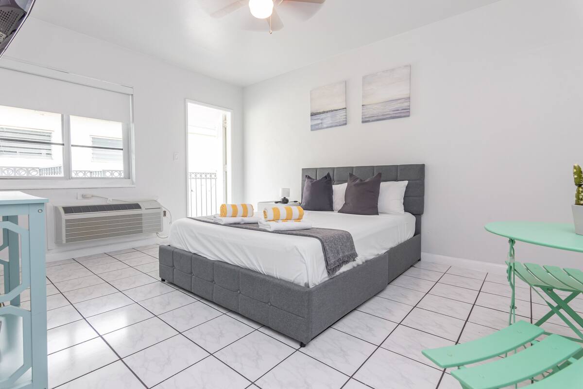 A modern bedroom with a grey upholstered bed, white bedding, and a green table with chairs.