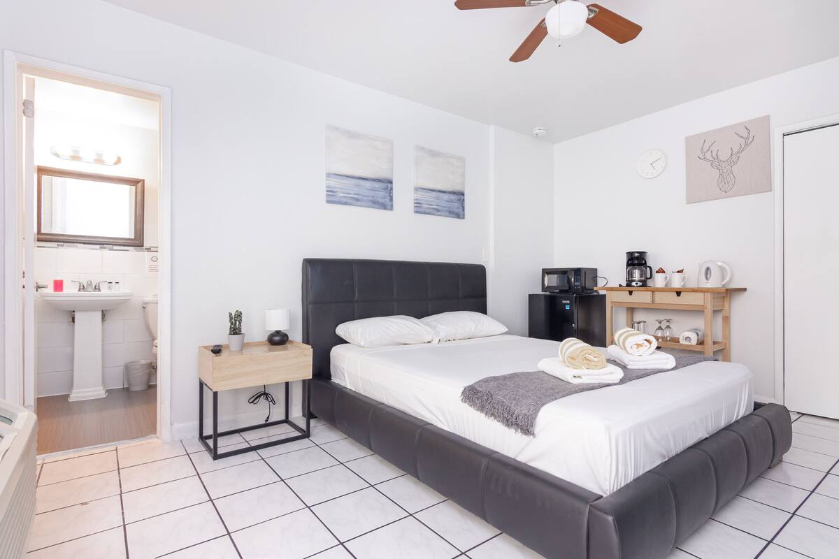 A modern bedroom with a large bed, bedside table, and a small desk. There is a bathroom with a sink and mirror.