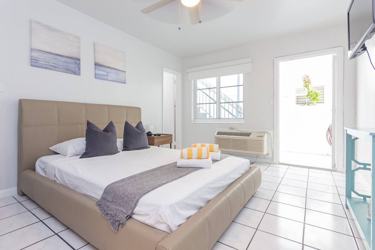 A bedroom with a large bed, a ceiling fan, a window with a view of a building, and a door leading outside.