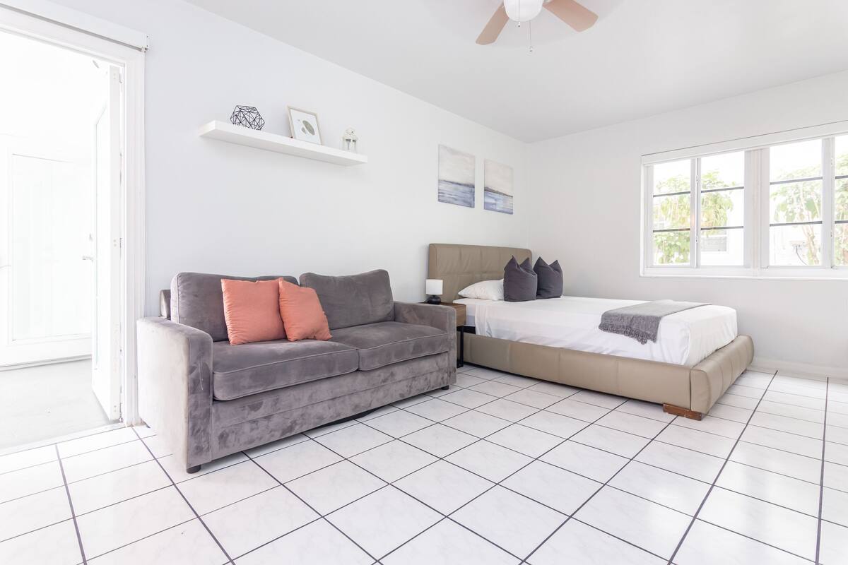 A modern bedroom with a grey sofa, a bed with a beige headboard, and a window with a view of greenery.