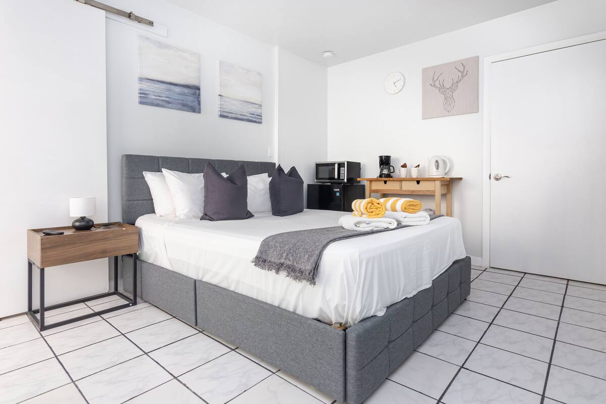 A modern bedroom with a grey upholstered bed, white bedding, a wooden bedside table, a microwave, a coffee maker, and a small shelf with towels.