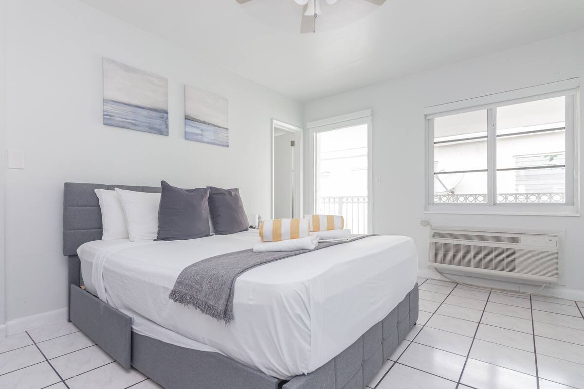A modern bedroom with a large bed, grey headboard, white bedding, and a window with a view.