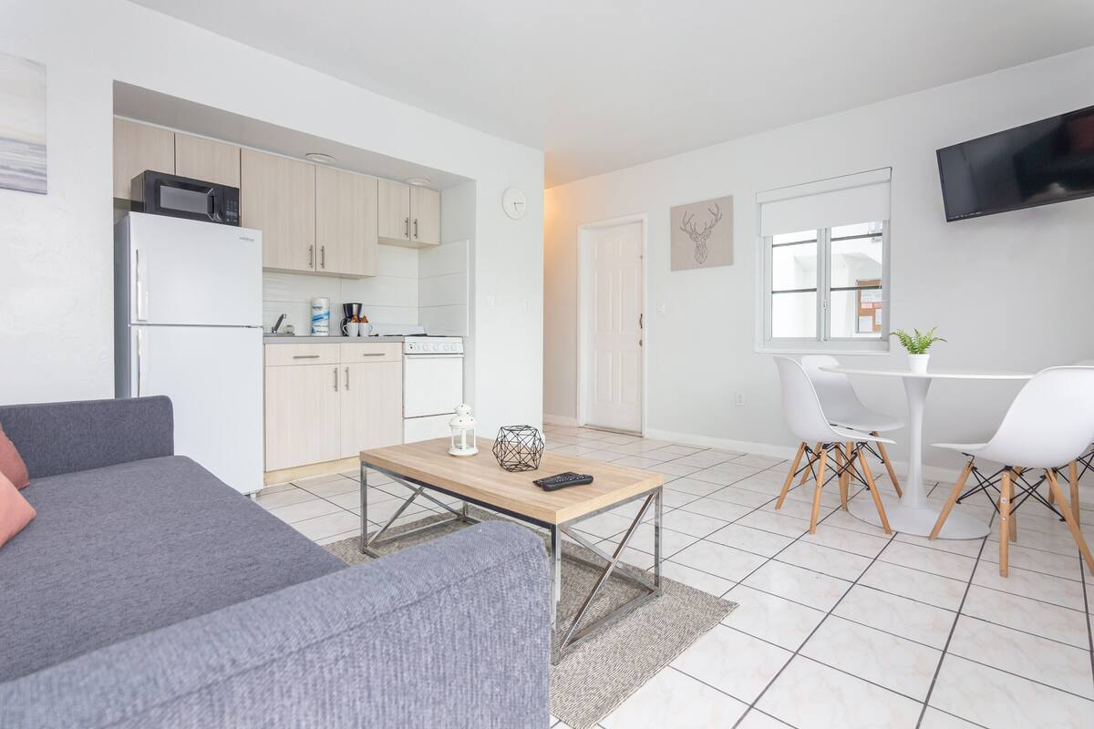 A modern living room with a grey sofa, a wooden coffee table, and a dining area with a white table and chairs.