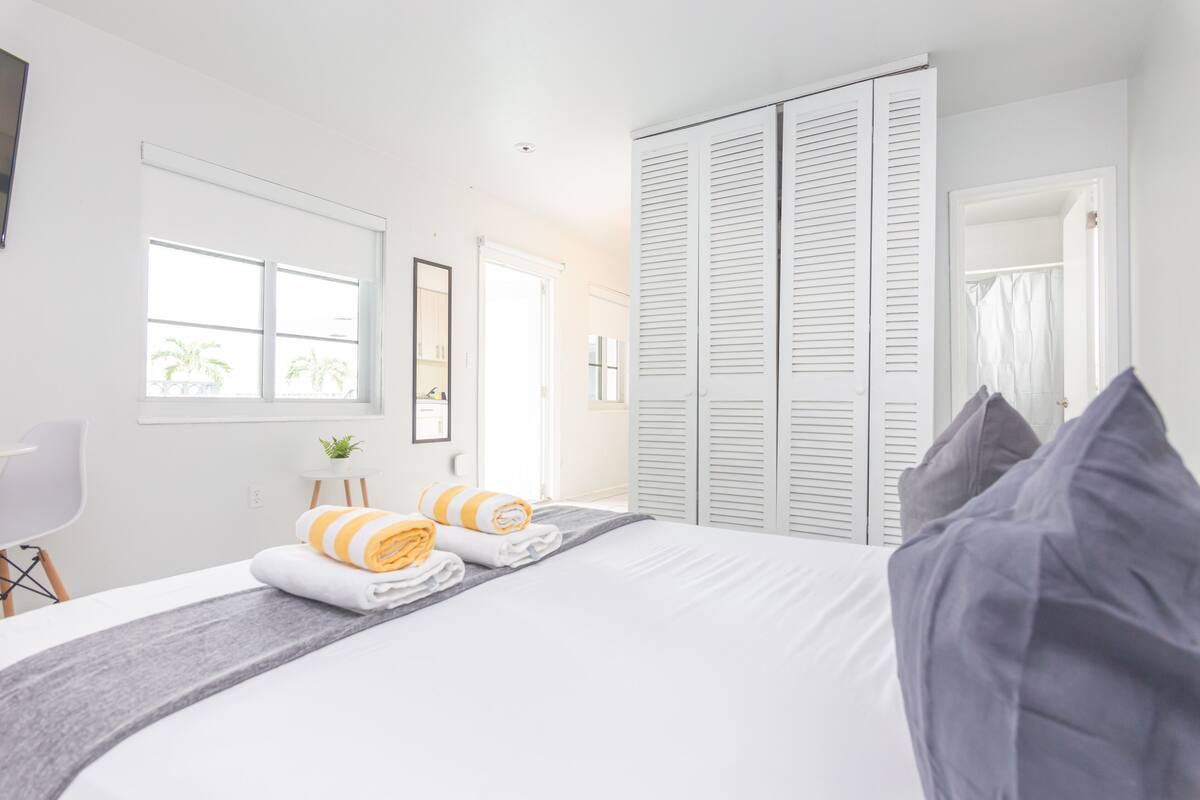 A hotel room with a bed, a desk, a chair, a window with a view of palm trees, and a wardrobe with sliding doors.