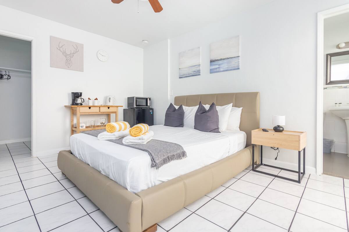 A modern hotel room with a large bed, a wooden bedside table, a microwave, and a small kitchen area.