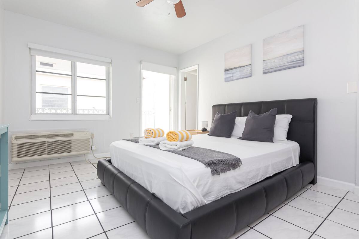 A modern bedroom with a large bed, white walls, and a view of the outdoors through a window.